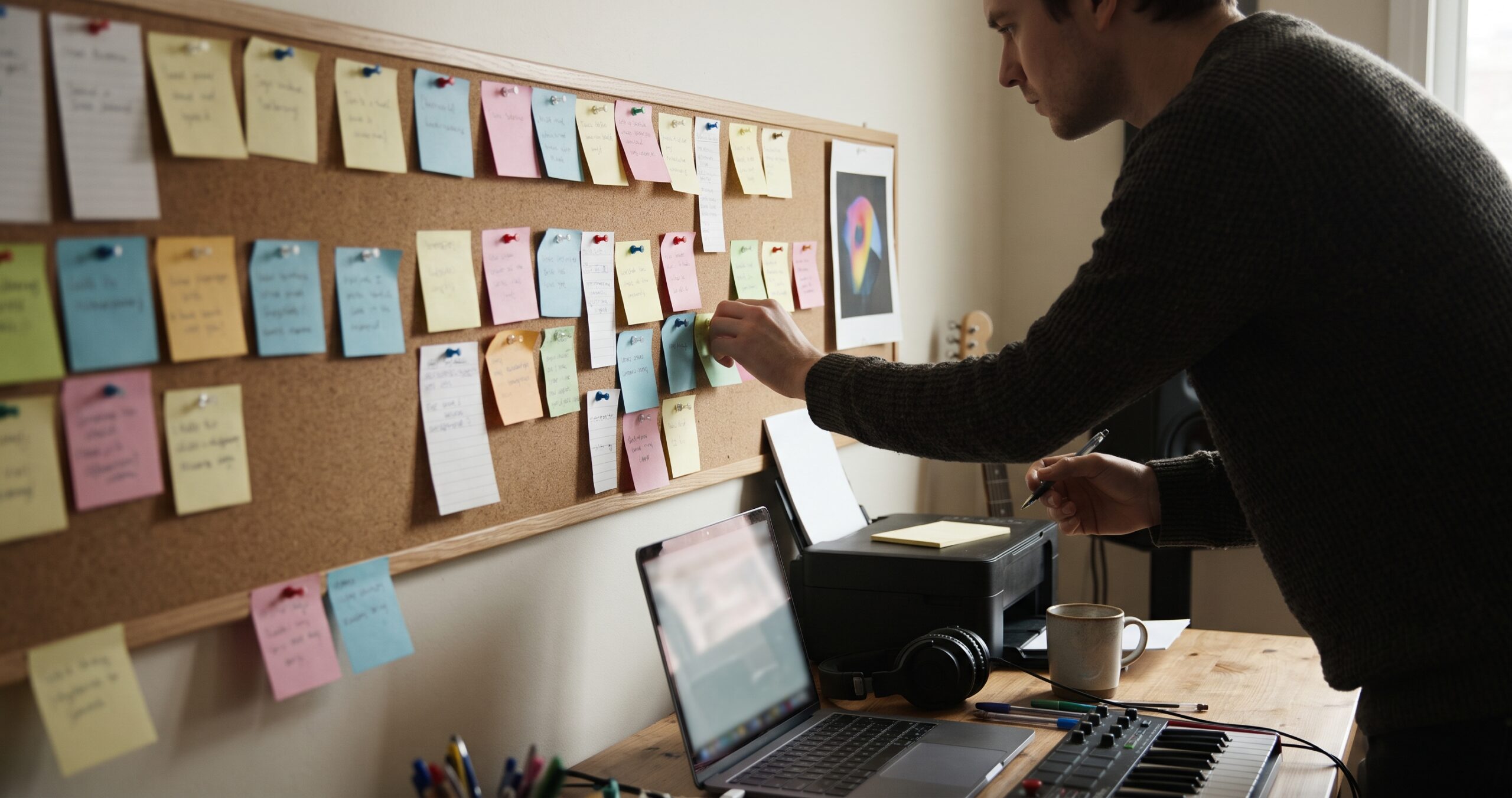 A musician pinning a sticky note to a long timeline of notes on a corkboard, with a cover-art print at the end and a laptop and headphones on the desk below.